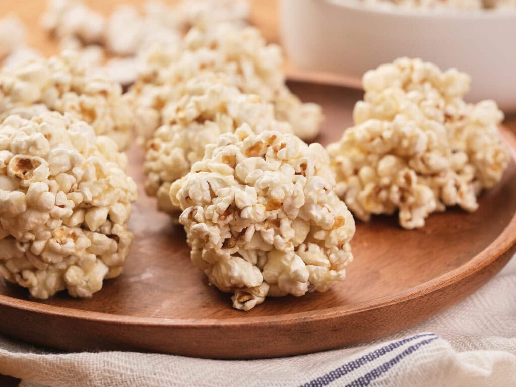 Several popcorn balls are arranged on a wooden plate, with a white bowl and more popcorn balls visible in the background.