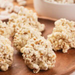 Several popcorn balls are arranged on a wooden plate, with a white bowl containing more popcorn in the background.