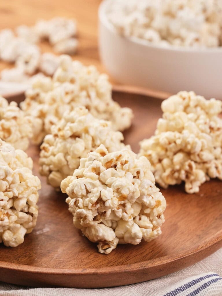 Several popcorn balls are arranged on a wooden plate, with a white bowl containing more popcorn in the background.