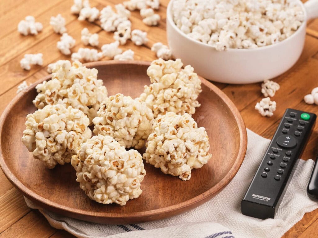 A wooden plate holds five popcorn balls, with a bowl of popcorn, scattered popcorn pieces, and a remote control on a wooden table.