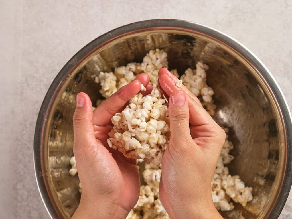Two hands are forming a ball of popcorn over a metal bowl filled with more popcorn.