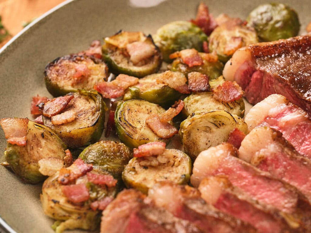 A plate with sliced steak and roasted Brussels sprouts topped with pieces of bacon.