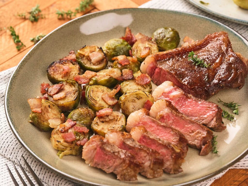 A plate of sliced medium-rare steak served with roasted Brussels sprouts and pieces of bacon, garnished with herbs.