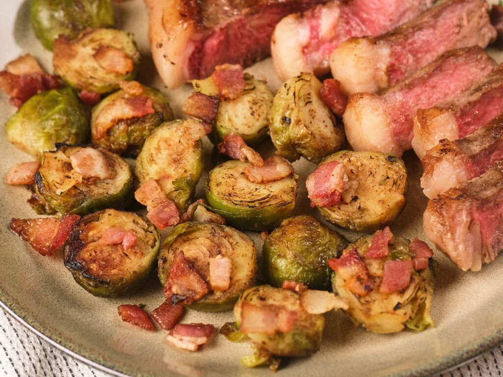 A plate with sliced steak and roasted Brussels sprouts topped with small pieces of bacon.