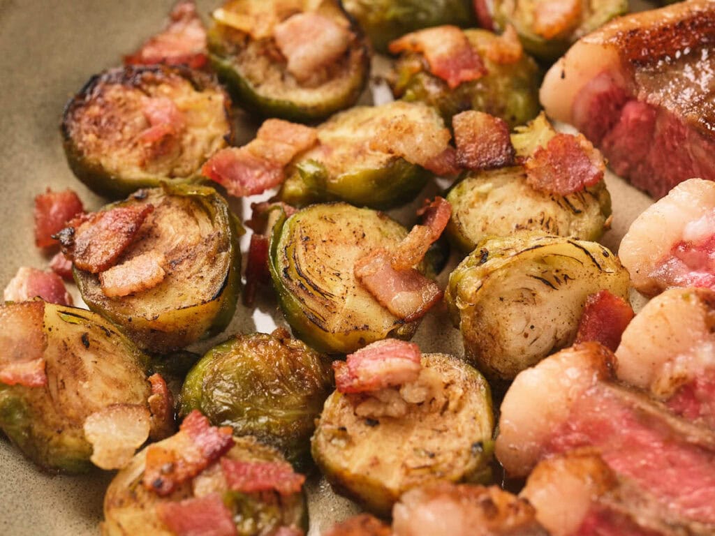 Close-up of roasted Brussels sprouts topped with small pieces of crispy bacon and slices of seared meat on a plate.