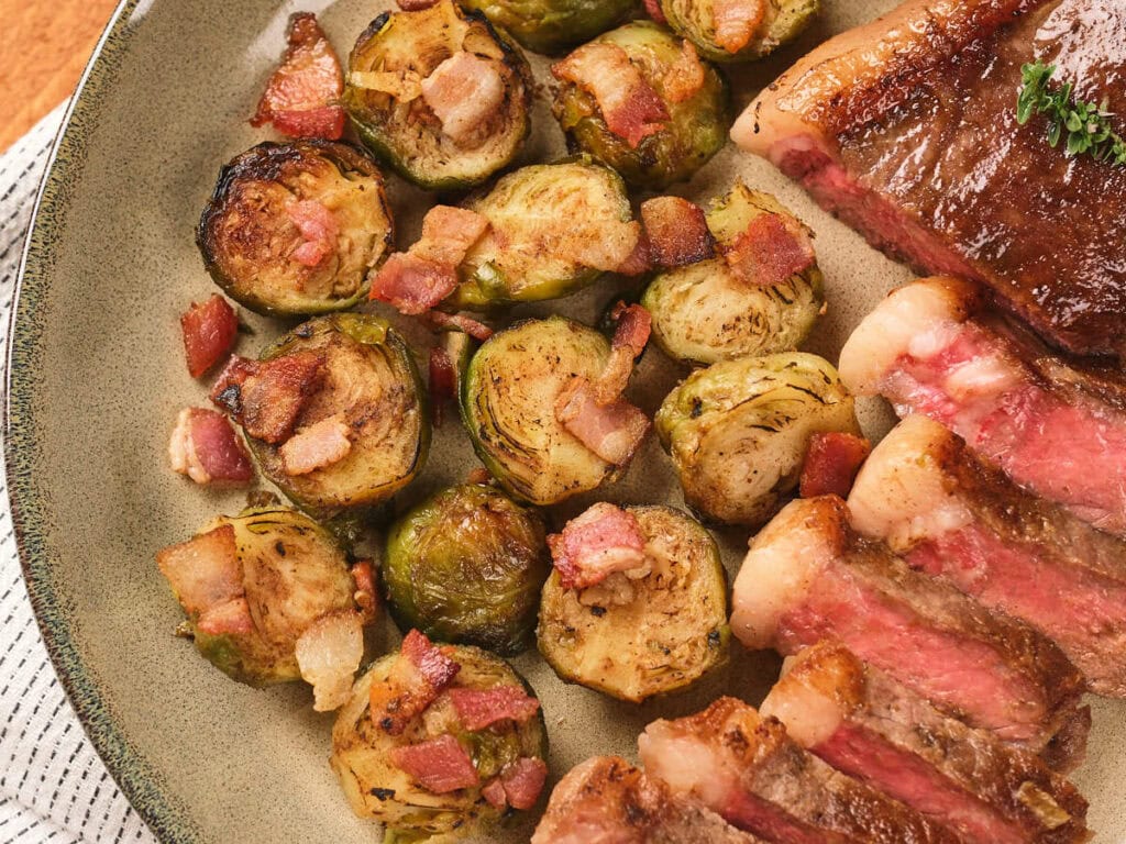 A plate with roasted Brussels sprouts topped with bacon pieces next to slices of medium-rare steak.