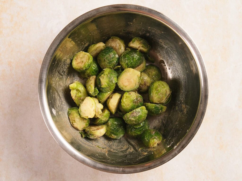 Halved Brussels sprouts coated in seasoning are placed in a stainless steel mixing bowl on a light-colored surface.