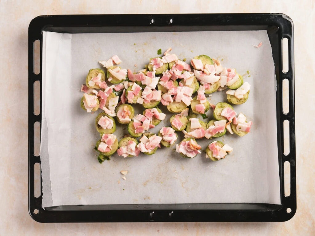 A baking tray lined with parchment paper holds sliced Brussel sprouts topped with diced bacon, ready to be cooked.