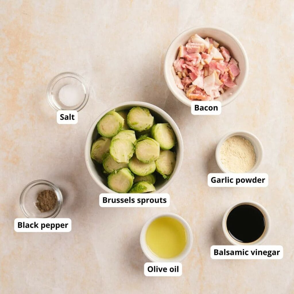 Top-down view of labeled ingredients in small bowls: Brussels sprouts, bacon, garlic powder, balsamic vinegar, olive oil, salt, and black pepper.