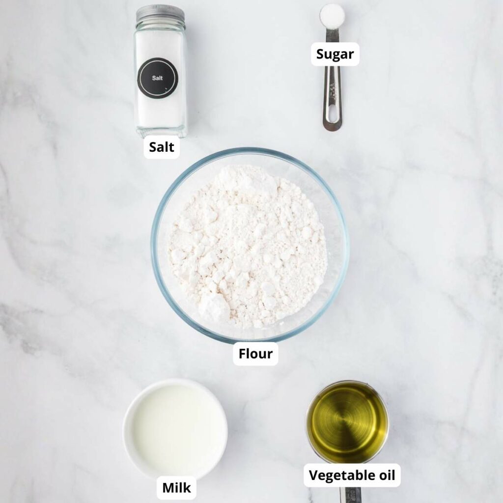 A bowl of flour, a cup of milk, a cup of vegetable oil, a jar of salt, and a measuring spoon of sugar are arranged on a white surface, each labeled.