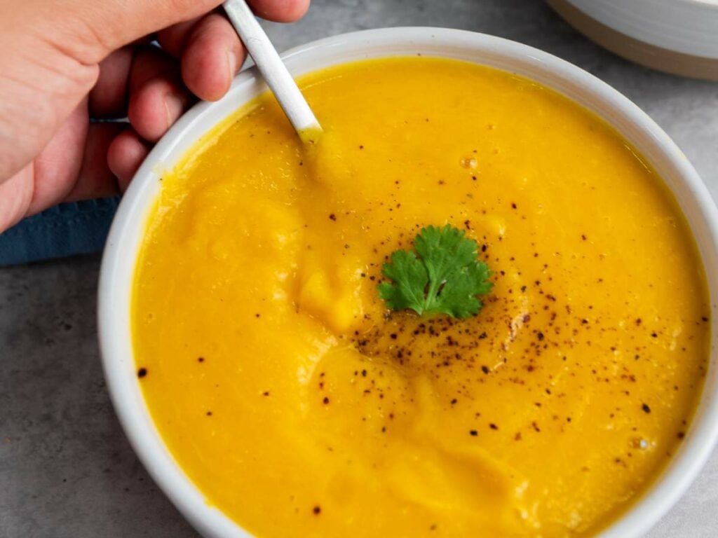 A bowl of creamy butternut squash soup garnished with black pepper and a cilantro leaf, with a hand holding a spoon.