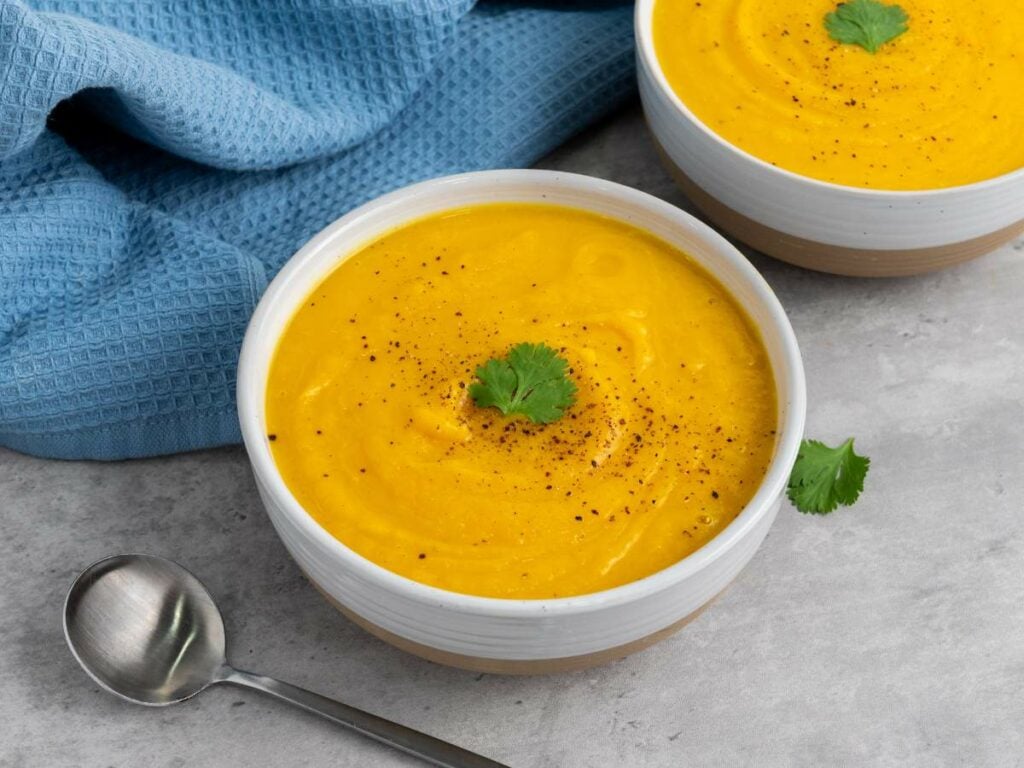 Two bowls of butternut squash soup topped with black pepper and a cilantro leaf, placed on a gray surface next to a spoon and a blue textured cloth.