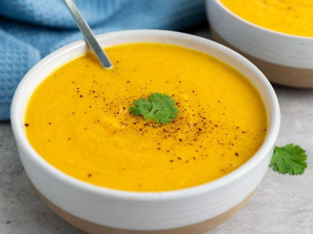 A bowl of creamy squash soup garnished with a sprig of cilantro and sprinkled with black pepper, with a spoon resting inside.