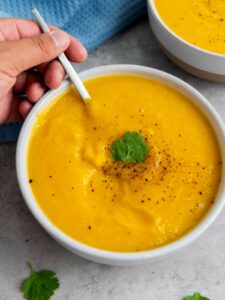 A hand holds a spoon in a bowl of butternut squash soup garnished with black pepper and a sprig of cilantro, with another bowl and a blue cloth nearby.