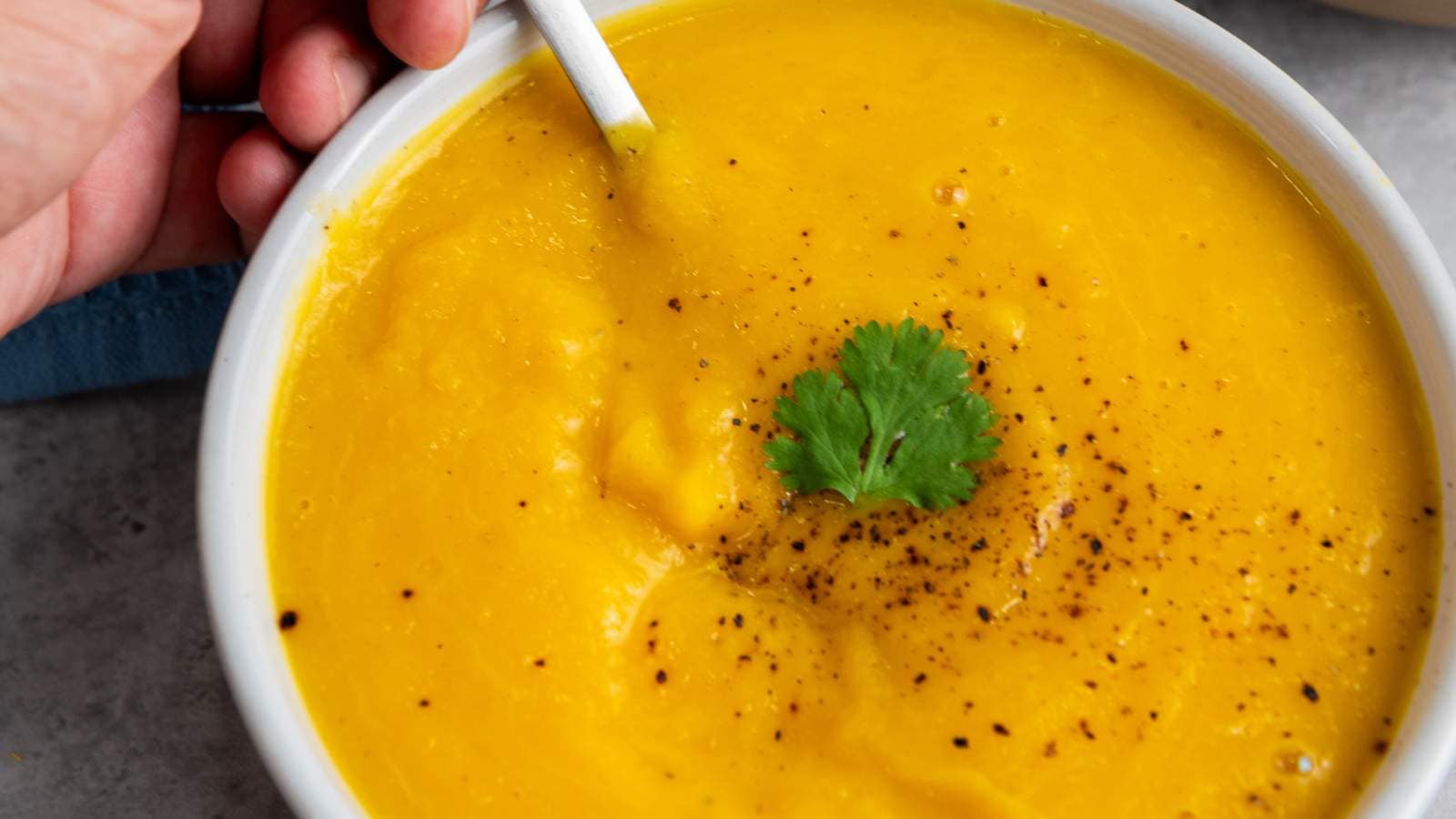 A bowl of butternut squash soup garnished with ground black pepper and a sprig of cilantro, with a hand holding the bowl and a spoon inside.