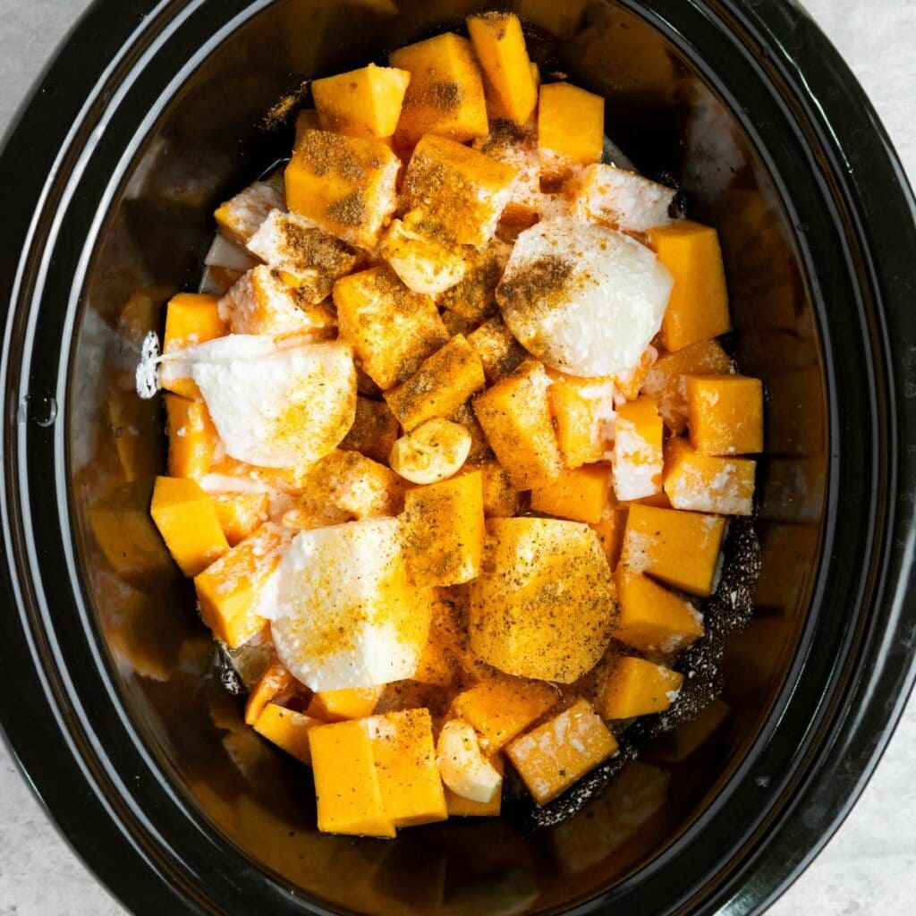 Cubed butternut squash, sliced onions, and seasonings are arranged inside a black slow cooker, ready for cooking.
