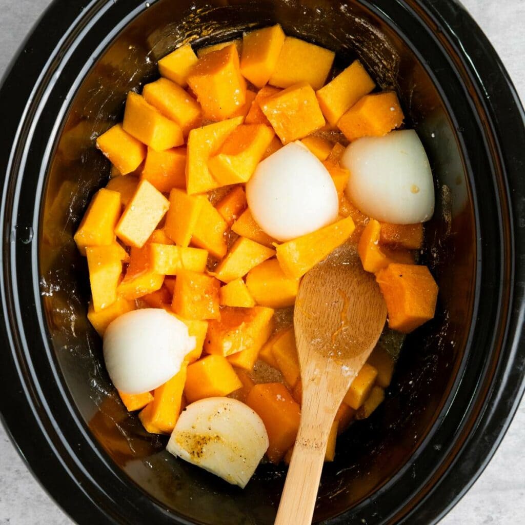 Cubed butternut squash and quartered onions in a slow cooker with a wooden spoon resting inside.