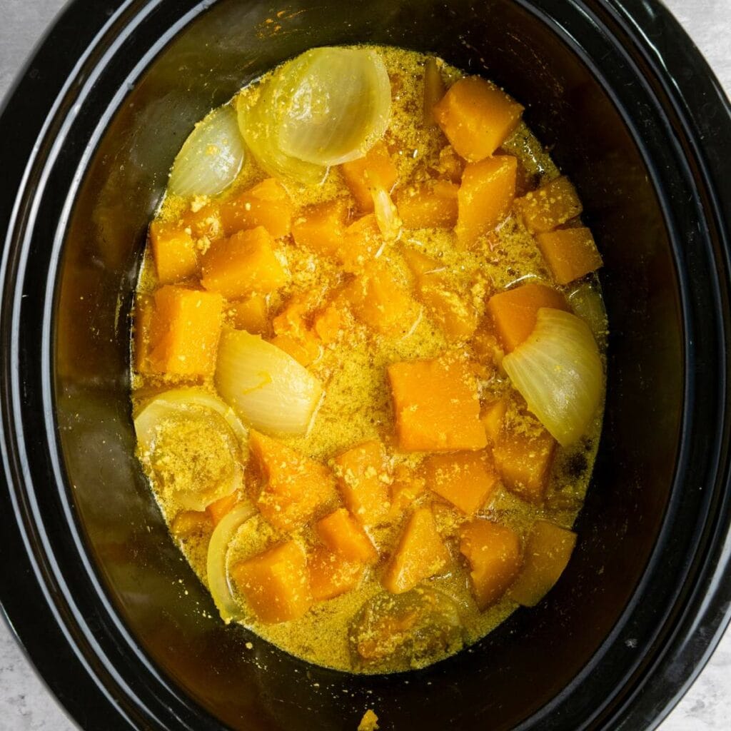 Chopped butternut squash and onion pieces in a slow cooker with broth and spices, ready to be cooked.