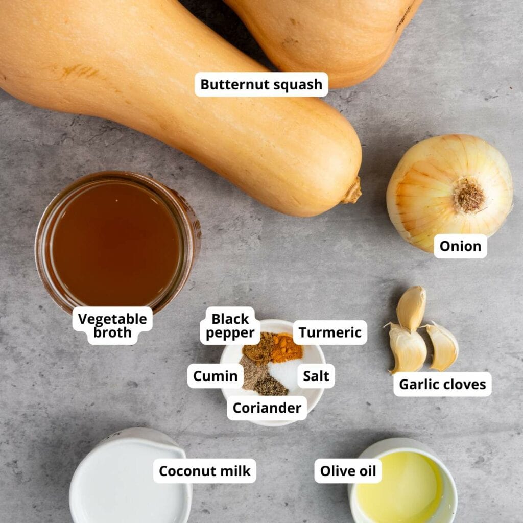 Ingredients for a soup arranged on a gray surface: butternut squash, onion, garlic, vegetable broth, coconut milk, olive oil, and assorted spices with labels.