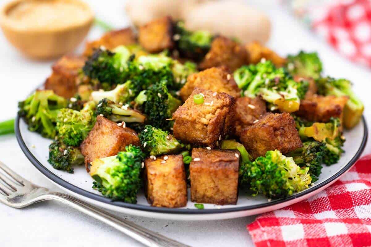 A plate of cubed tofu and broccoli tossed in sauce, garnished with sesame seeds, sits on a table next to a fork and a red checkered napkin.