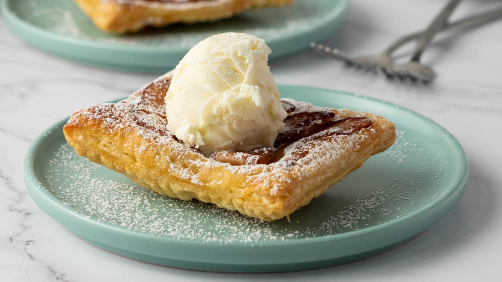 A square piece of apple tart topped with a scoop of vanilla ice cream sits on a light blue plate, dusted with powdered sugar.