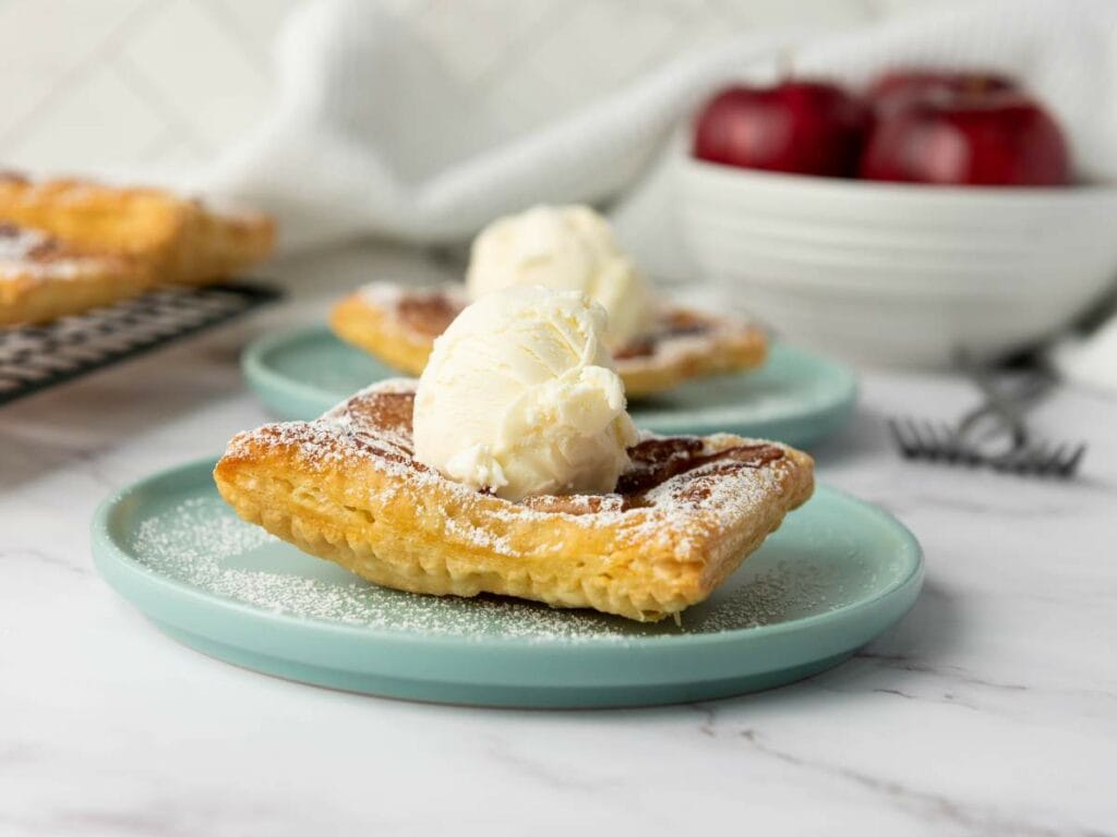 A square apple tart topped with a scoop of vanilla ice cream sits on a light blue plate, with more pastries and apples visible in the background.