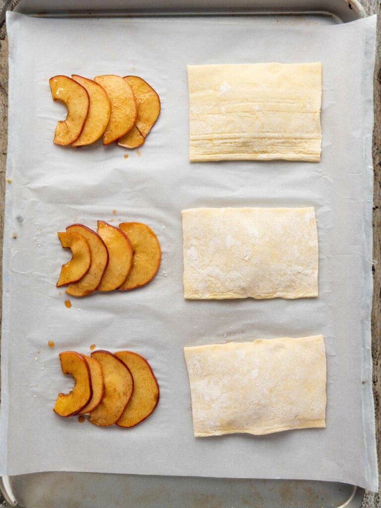 A baking sheet lined with parchment paper holds three rows of apple slices and three rectangles of uncooked puff pastry.