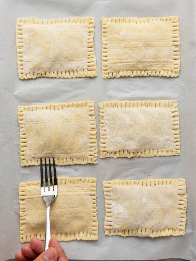 Six rectangular pieces of uncooked pastry with crimped edges on parchment paper; a hand presses a fork to seal one of the pastries.