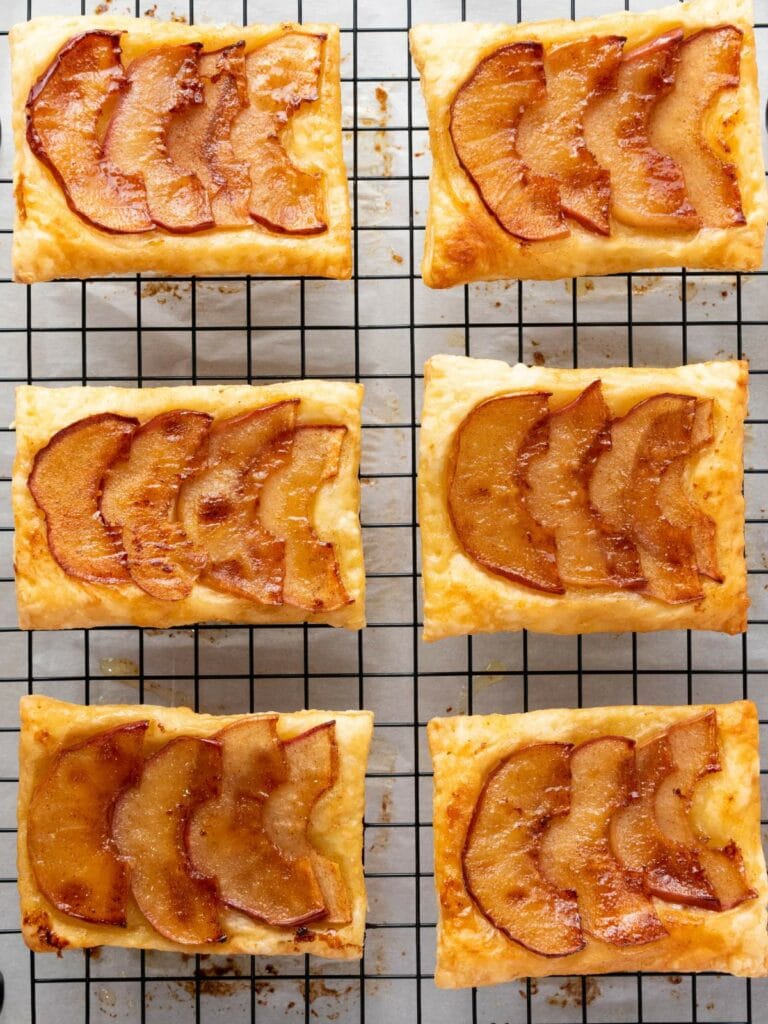 Six rectangular apple pastries are cooling on a wire rack, each topped with thinly sliced, baked apple arranged in a fan pattern.