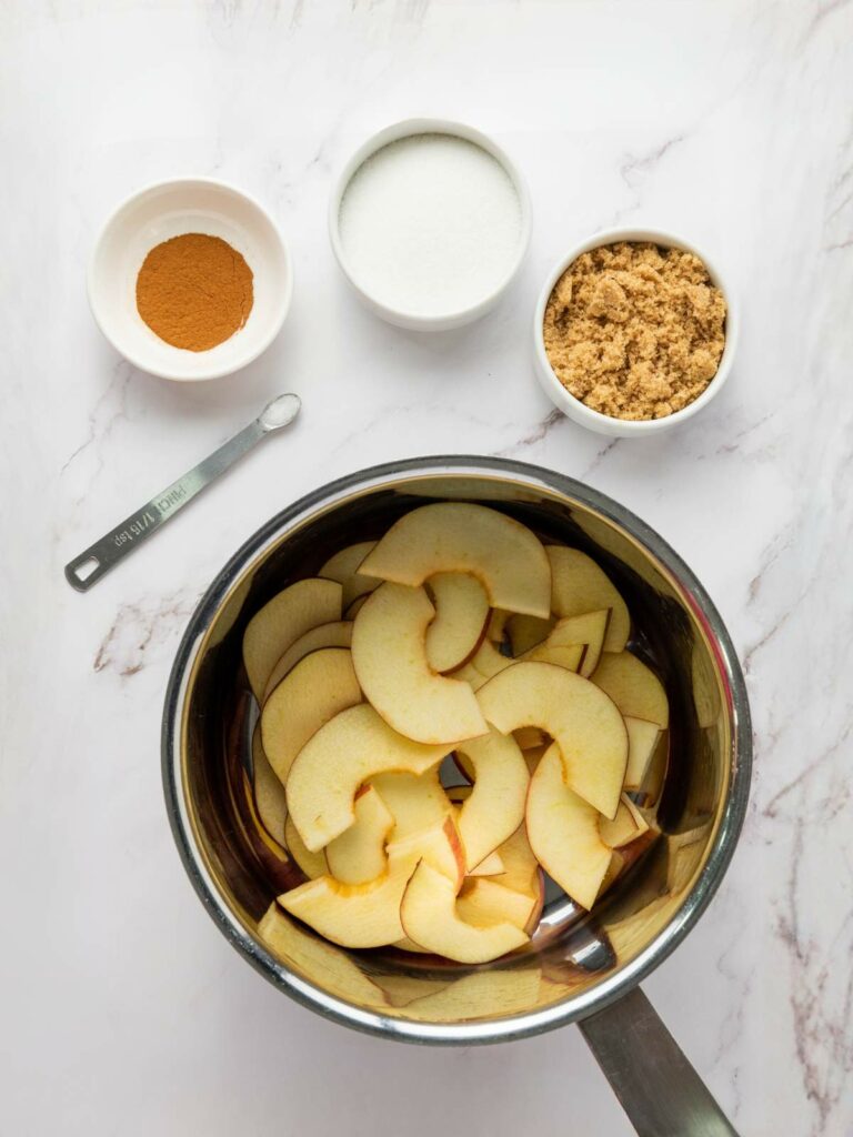 A saucepan with sliced apples, next to bowls of cinnamon, white sugar, and brown sugar, plus a measuring spoon, on a white surface.