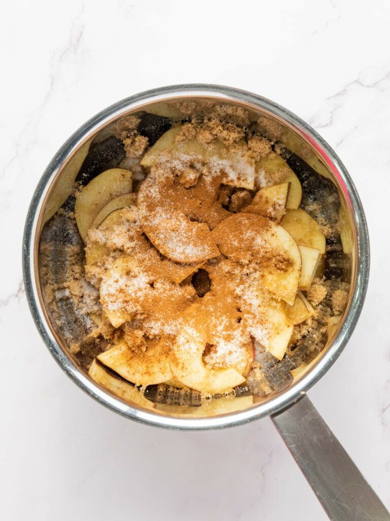 Sliced apples in a saucepan topped with brown sugar, granulated sugar, and ground cinnamon, ready for cooking.