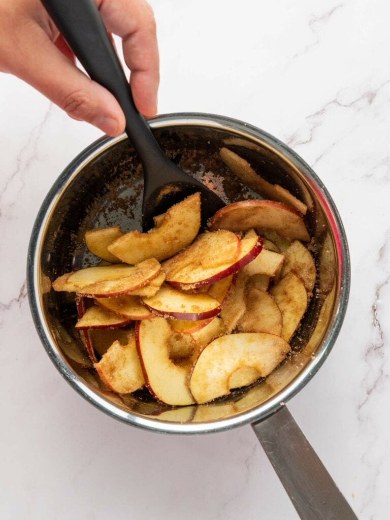 A hand stirs sliced apples with cinnamon and sugar in a saucepan using a black spoon on a white marble surface.