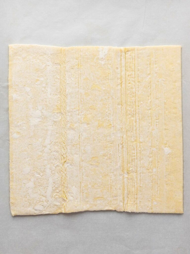 A square sheet of raw puff pastry dough is laid flat on a light-colored surface.
