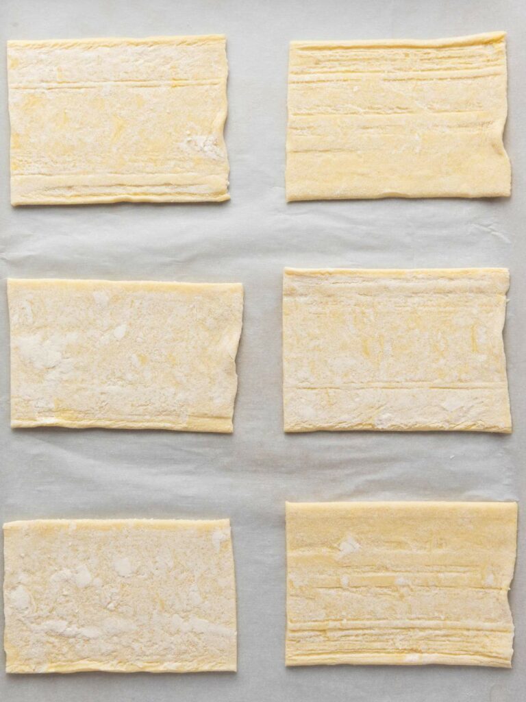 Six rectangular pieces of puff pastry dough are arranged on a sheet of parchment paper.
