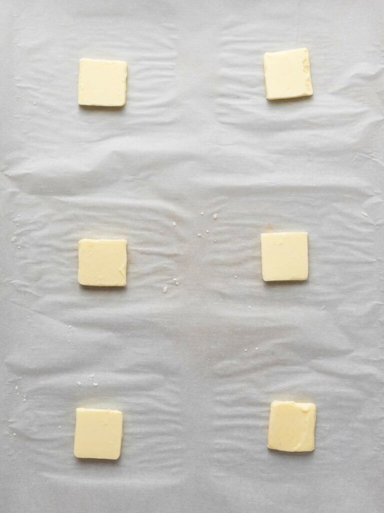 Six slices of butter are evenly spaced on a sheet of parchment paper.