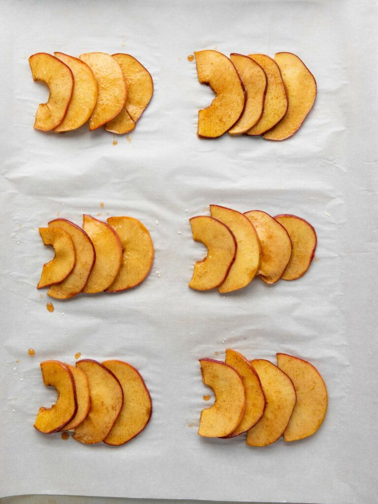 Six groups of thinly sliced, seasoned apple pieces are arranged in rows on a sheet of parchment paper.