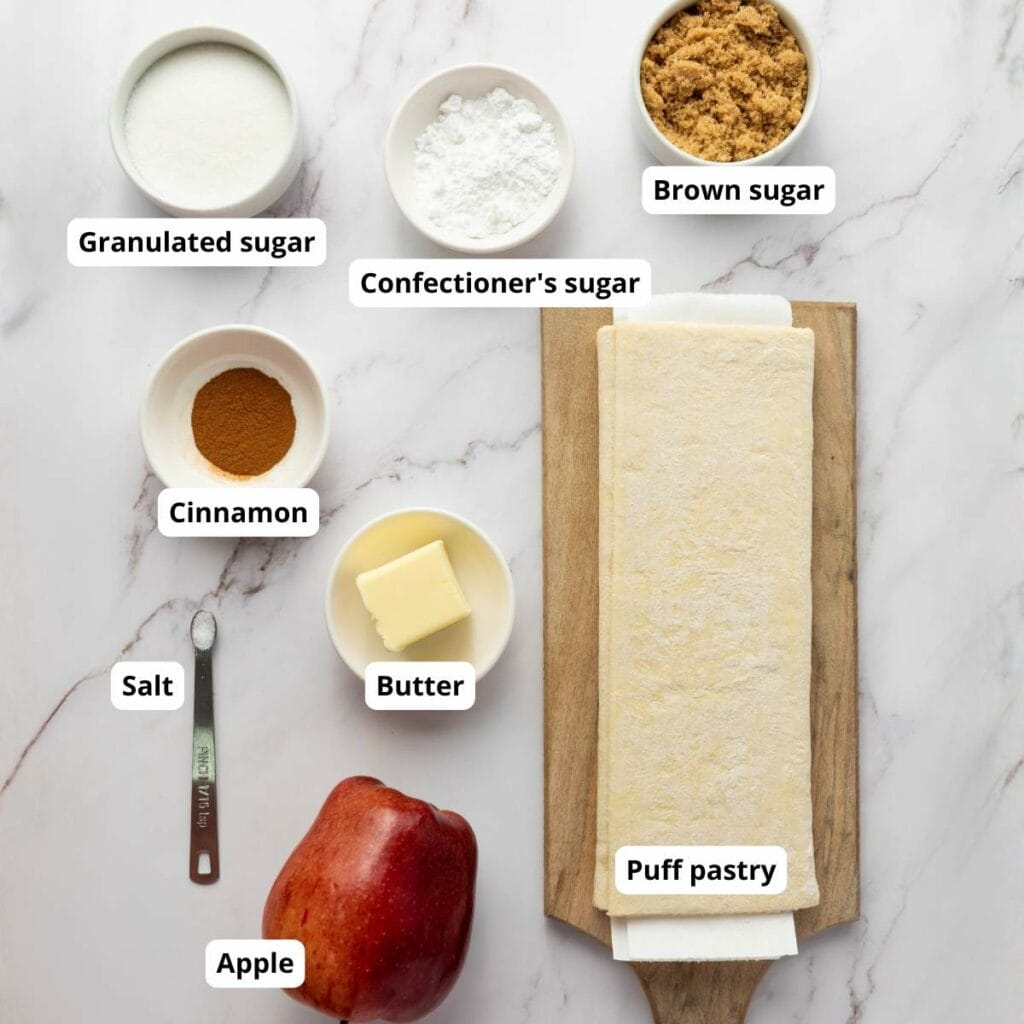 Ingredients on a marble surface: granulated sugar, confectioner's sugar, brown sugar, cinnamon, salt, butter, apple, and a sheet of puff pastry.