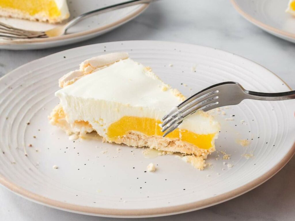 A slice of lemon meringue pie on a white plate with a fork cutting into it; two other plates are partially visible in the background.