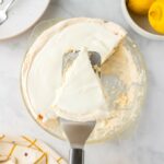 A hand holds a spatula lifting a slice of lemon angel pie with a creamy topping from a glass pie dish; lemons and plates are nearby on a marble surface.