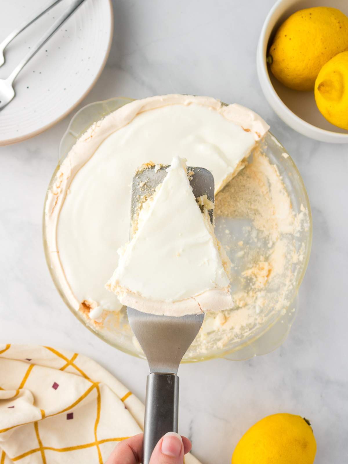 A hand holds a spatula lifting a slice of lemon angel pie with a creamy topping from a glass pie dish; lemons and plates are nearby on a marble surface.