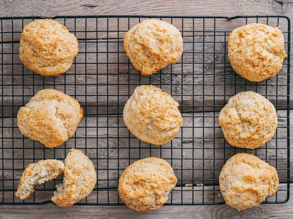 Nine golden-brown drop biscuits, one partially eaten, rest on a black wire cooling rack set on a wooden surface.
