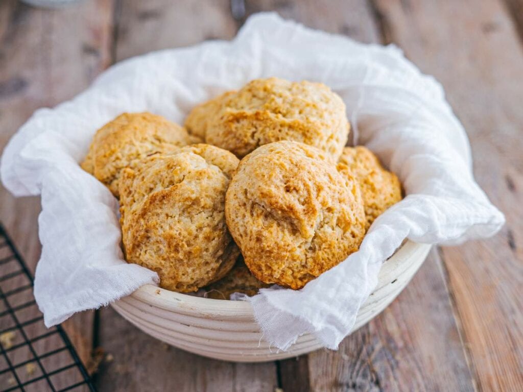 A bowl lined with a white cloth holds several drop biscuits on a rustic wooden table.