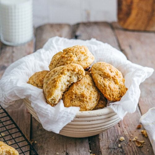A ceramic bowl lined with a white cloth holds several golden scones on a wooden table, with a glass of milk.