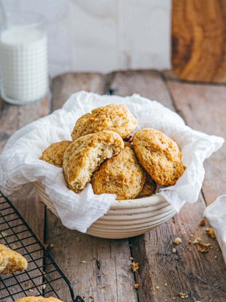 A ceramic bowl lined with a white cloth holds several golden scones on a wooden table, with a glass of milk.