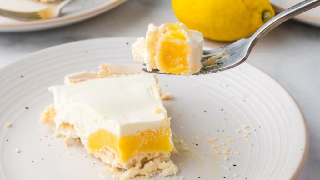 A piece of lemon dessert with a creamy white layer and crumbly crust on a plate, with a fork holding a bite and a lemon in the background.