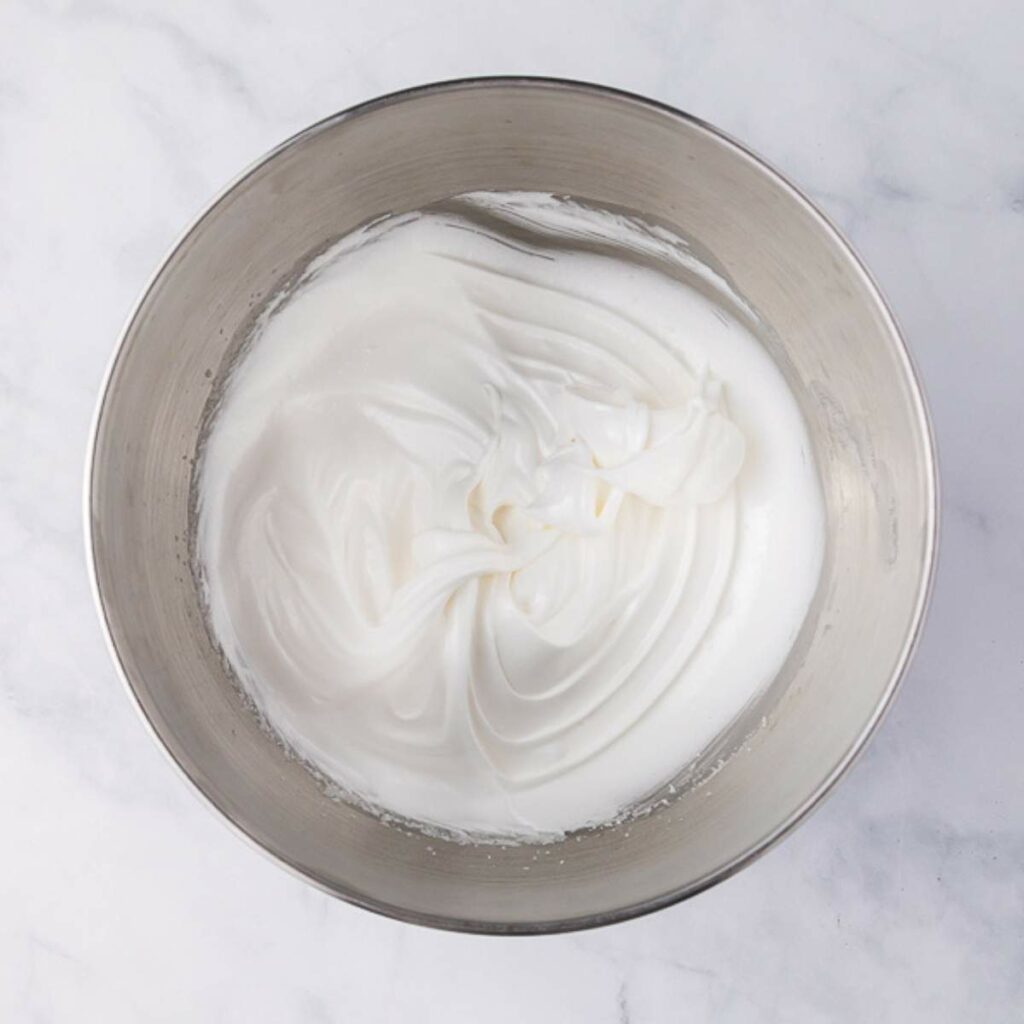 A stainless steel mixing bowl filled with whipped egg whites forming soft peaks, placed on a white marble surface.