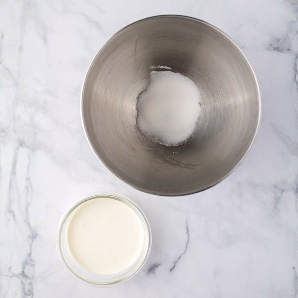 A metal mixing bowl containing sugar and a glass bowl filled with cream are placed on a white marble surface.