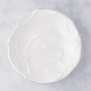 A round dish filled with a smooth layer of white whipped topping or meringue, viewed from above on a light-colored surface.