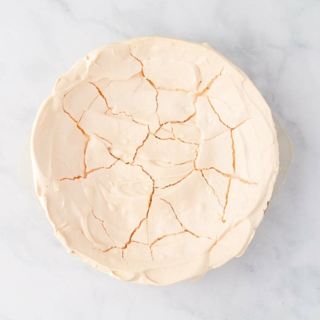 A round baked meringue shell with a cracked surface sits on a white marble background.