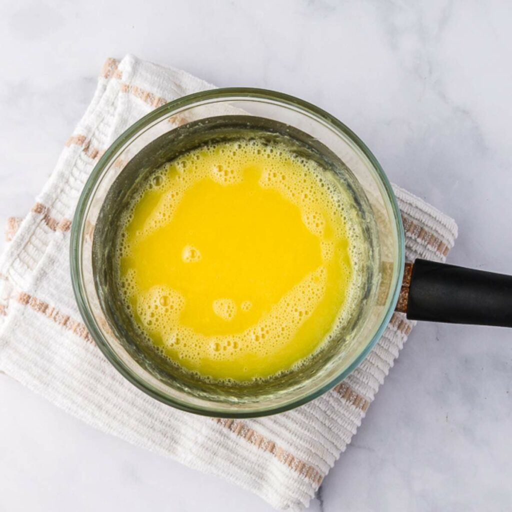 A saucepan filled with melted butter sits on a folded white kitchen towel with tan stripes, placed on a marble surface.